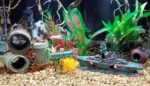 A detailed blue resin sunken ship decoration positioned within a vibrant aquarium, surrounded by green aquatic plants.